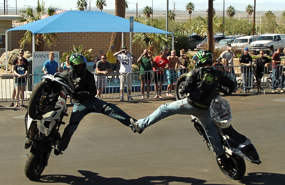 Dos motociclistas con casco ejecutan acrobacias durante una exhibición con público detrás de barreras.