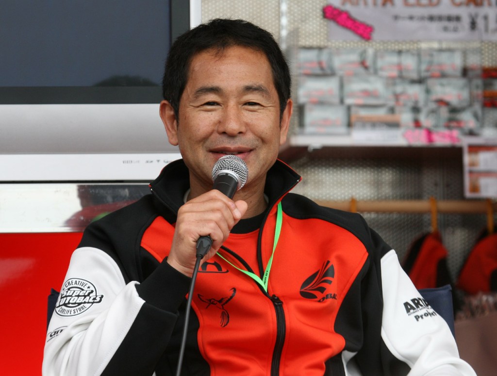 Keiichi Tsuchiya habla por un micrófono con indumentaria del equipo ARTA.
